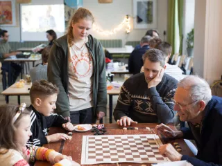 Vijf mensen rondom de tafel waarop een damspel ligt dat gespeeld wordt tussen een oudere meneer en een jonge jongen